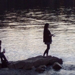 Two girls rest on a small jut of land in the middle of a lake, one fishing, one drinking a coffee.