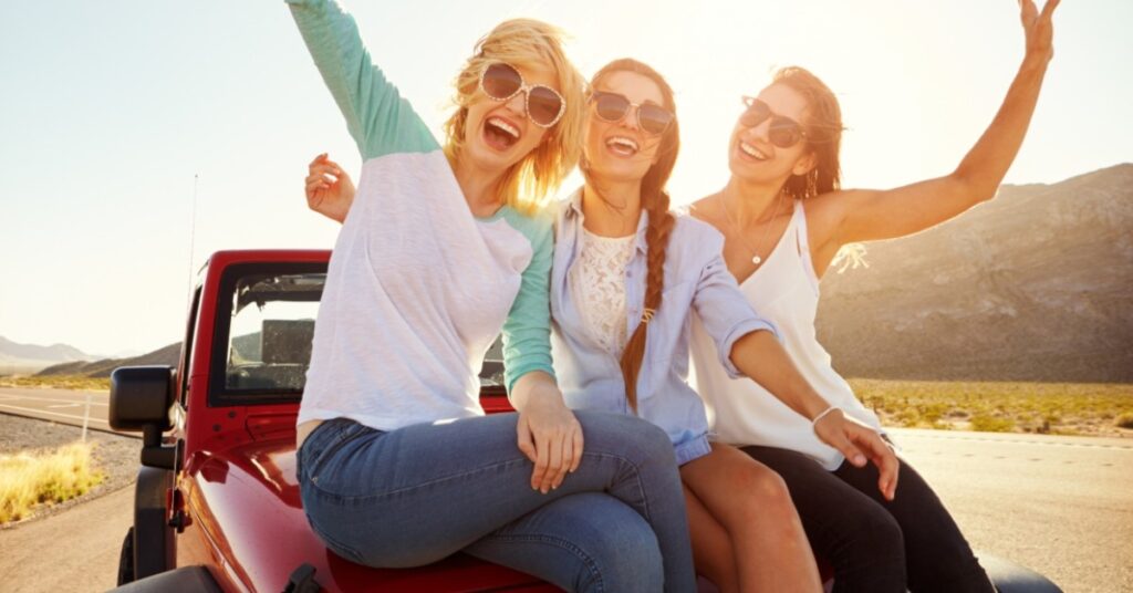 Three women sit on a red jeep as the sun sets behind them.