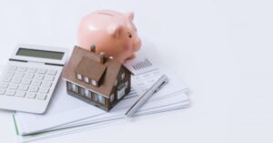 A piggy bank, model house and calculator rest on top of a packet of paper.